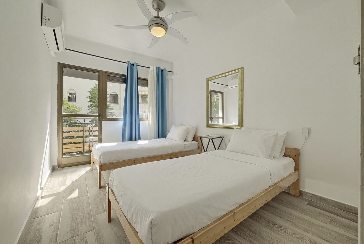 A bright bedroom with two twin beds, white linens, a ceiling fan, air conditioning, and a balcony visible through glass doors.