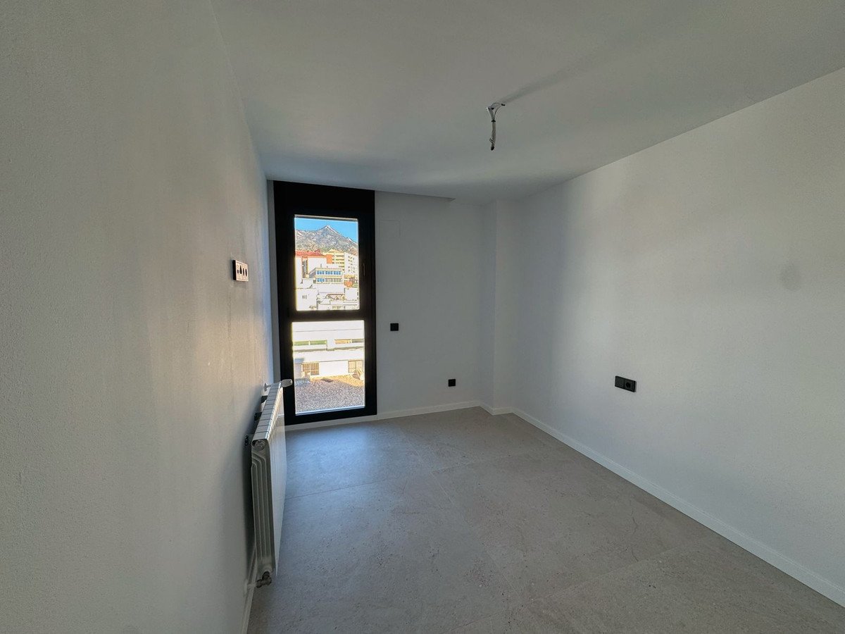 Empty room with a large window showing a mountain landscape and buildings. Features a radiator and light-colored floor.