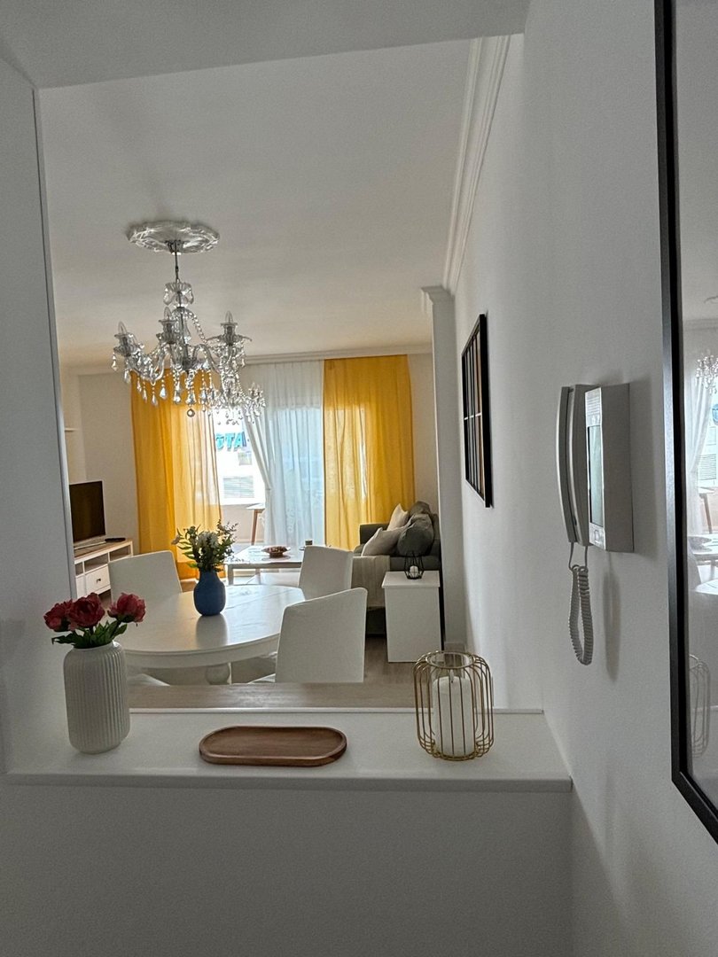 A dining area with a round table and chairs, and a living area with a sofa, illuminated by a crystal chandelier and yellow curtains.