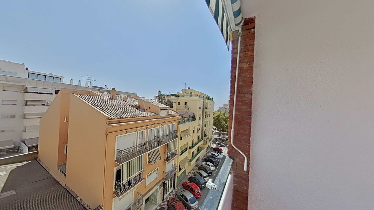 A view from a balcony overlooking a city street with parked cars and buildings under a clear blue sky.