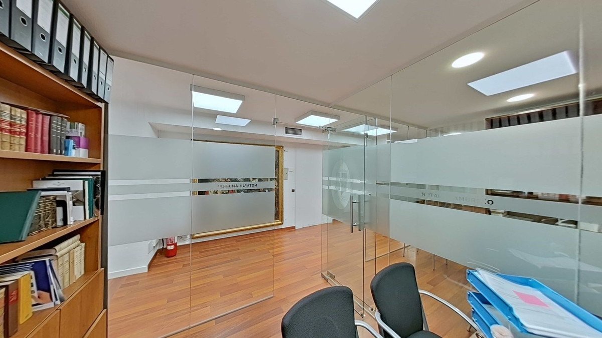 Modern office interior with glass walls, wooden floors, and a bookshelf filled with books. Features frosted privacy panels.