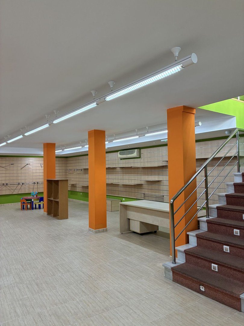 Empty commercial space with orange pillars, shelving on walls, a desk, and carpeted stairs with metal railings.