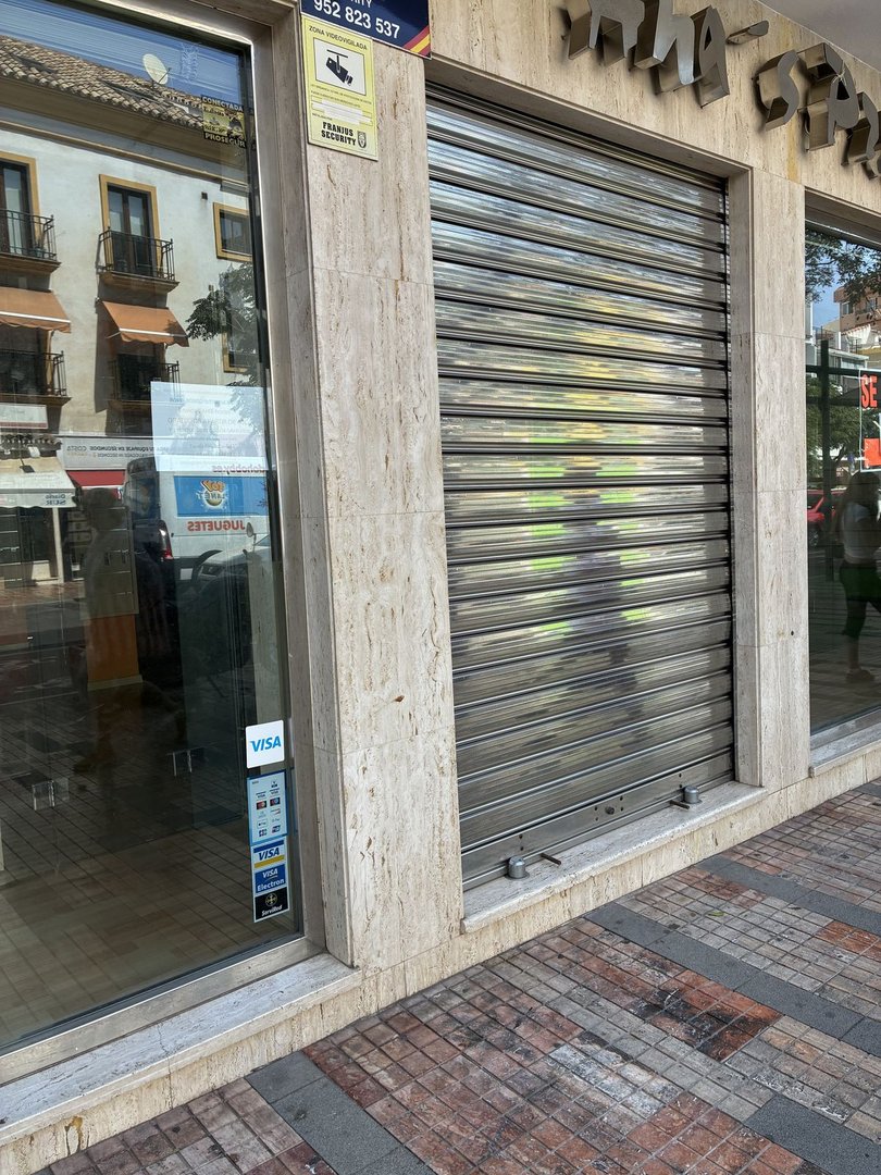 Street-level view of a commercial storefront with a closed roller shutter, marble accents, and Visa payment method stickers on the window.