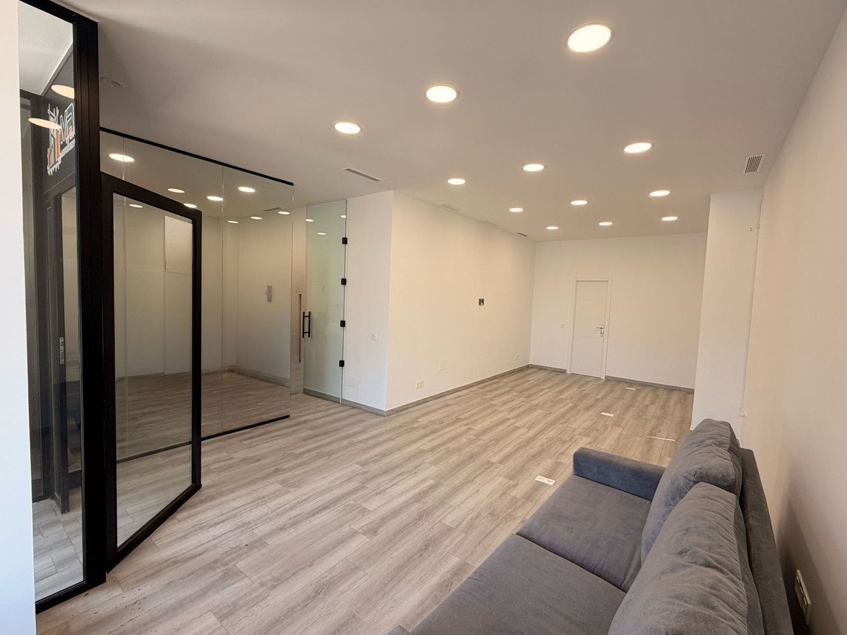 Modern office interior with glass walls, recessed lighting, wood-look flooring, and a grey sofa visible.
