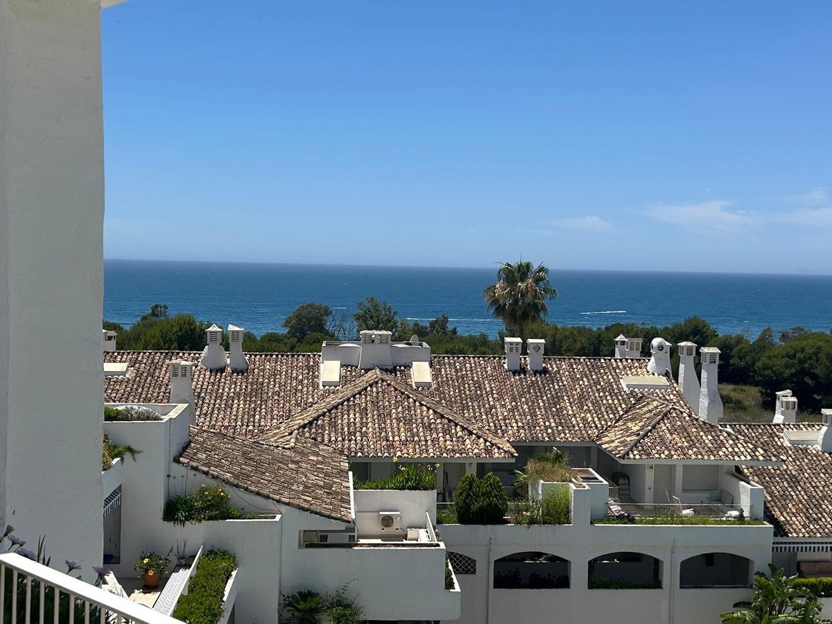 Sea view from a balcony overlooking white buildings with terracotta roofs and palm trees.
