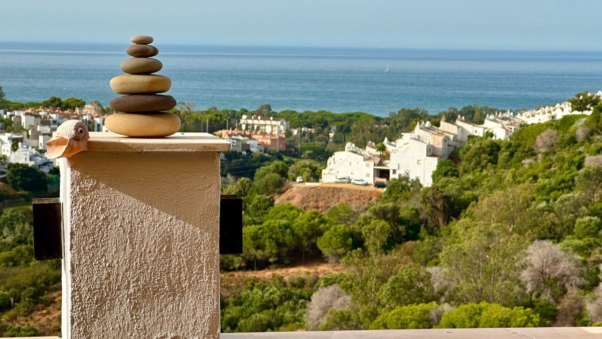 Balcony view with stacked stones and a decorative duck, overlooking white houses nestled among lush green trees and the blue sea.