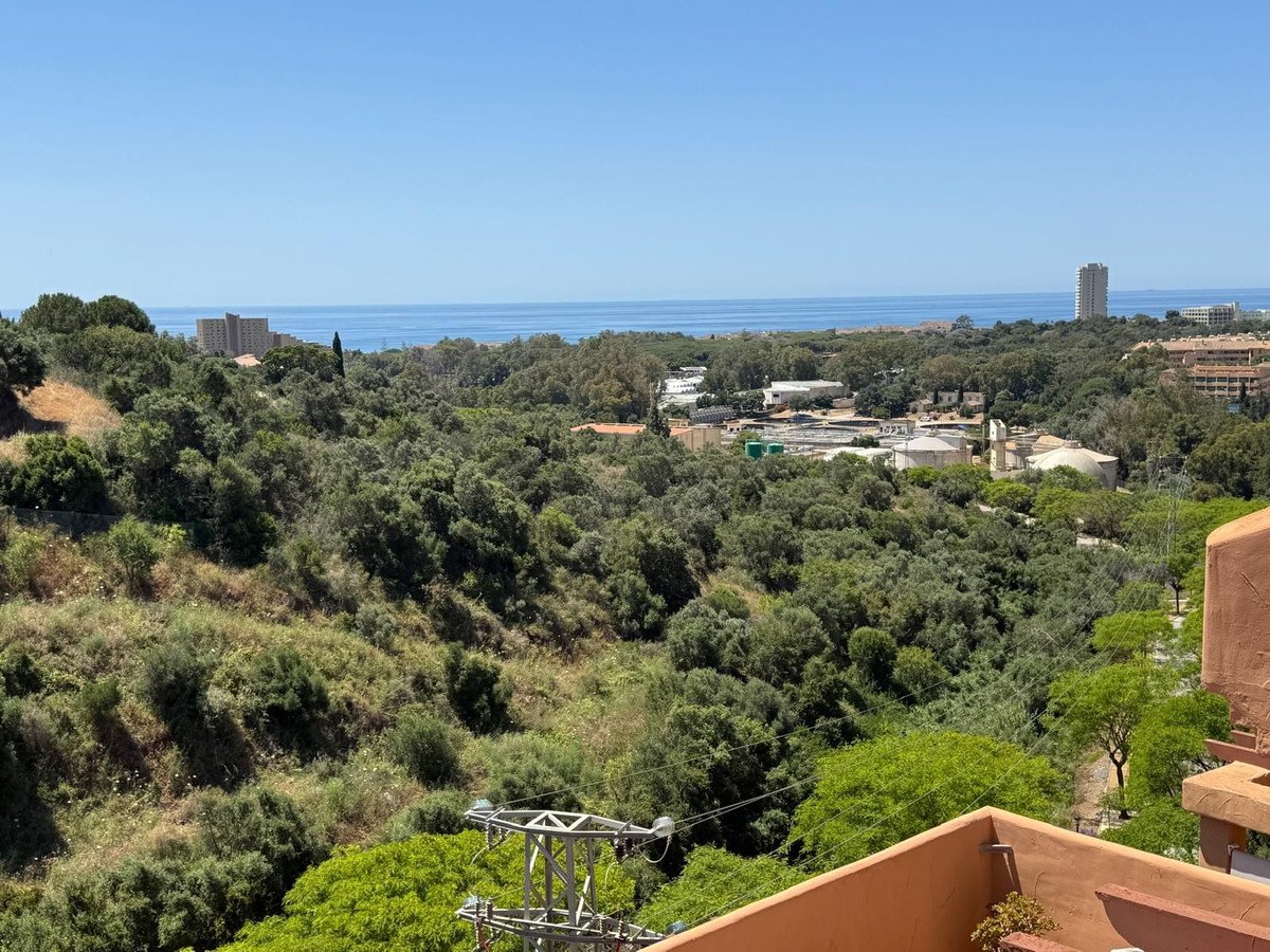 A scenic view from a balcony overlooking a lush green valley with buildings and the sea in the distance under a clear blue sky.