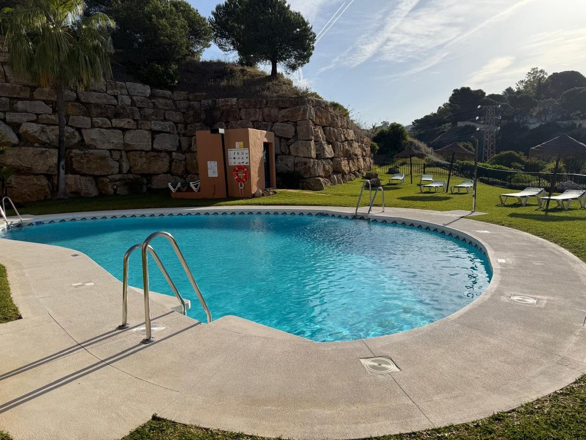 Outdoor swimming pool with sun loungers and parasols on a sunny day, surrounded by lush green grass and a stone wall.