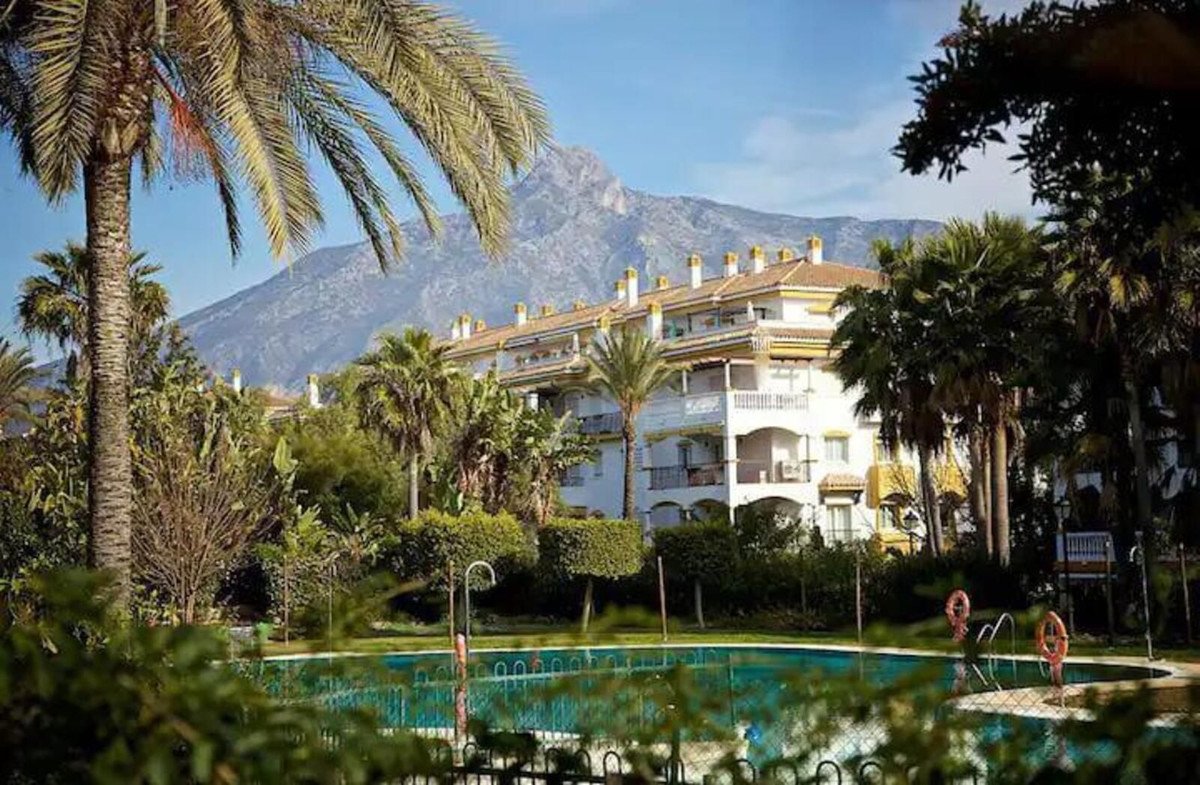 Apartment building with balconies and mountain backdrop, featuring a swimming pool and palm trees in the foreground.