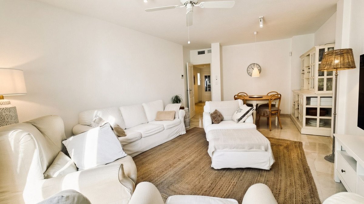 A bright living room with white sofas, a dining area with wooden chairs, and a decorative cabinet.
