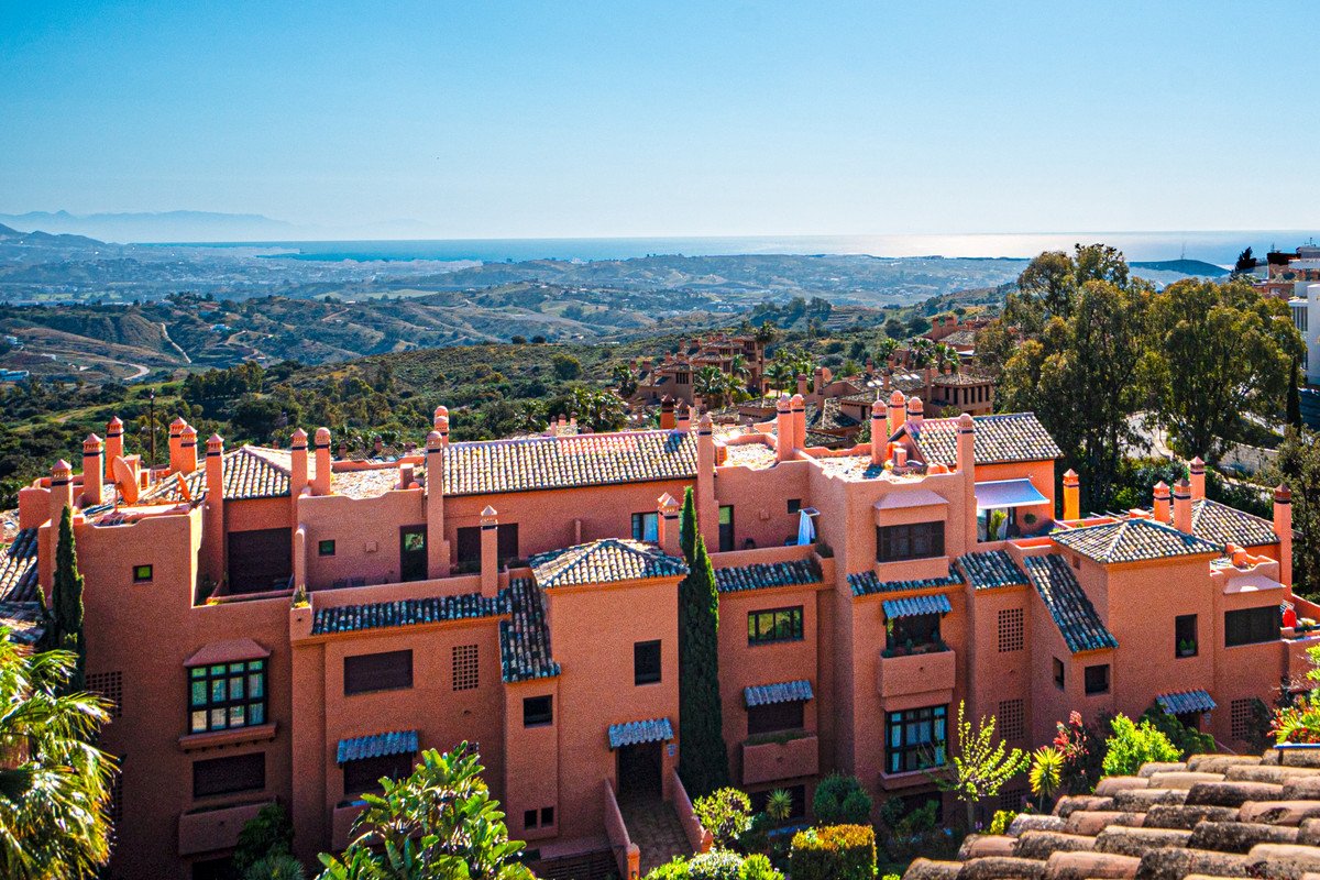 Terracotta apartment complex with tiled roofs, overlooking rolling hills and the sea under a clear blue sky.