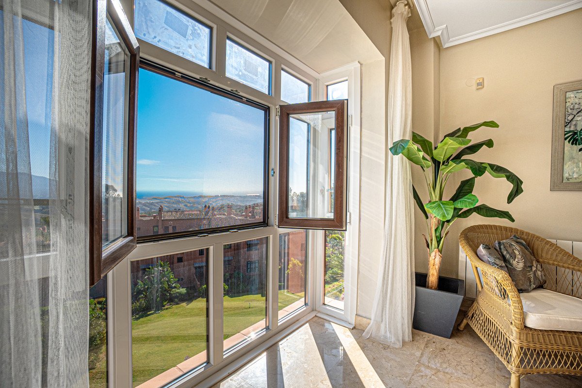 Sunlit balcony with large windows, wicker chair, potted plant, and panoramic sea view.