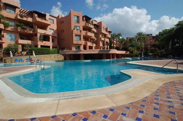 A wide shot of a large, blue swimming pool in front of a modern resort-style apartment building.