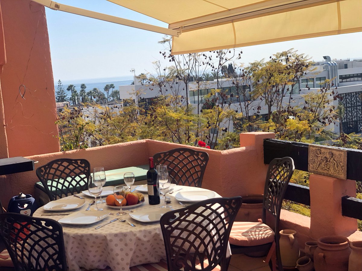 Set dining table on a terrace with a sea view, surrounded by trees and buildings.