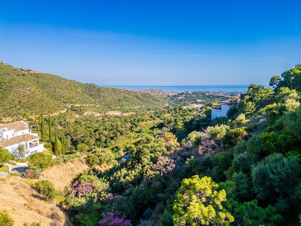 A white villa nestled on a green hillside with a distant view of the sea and coastline under a clear blue sky.