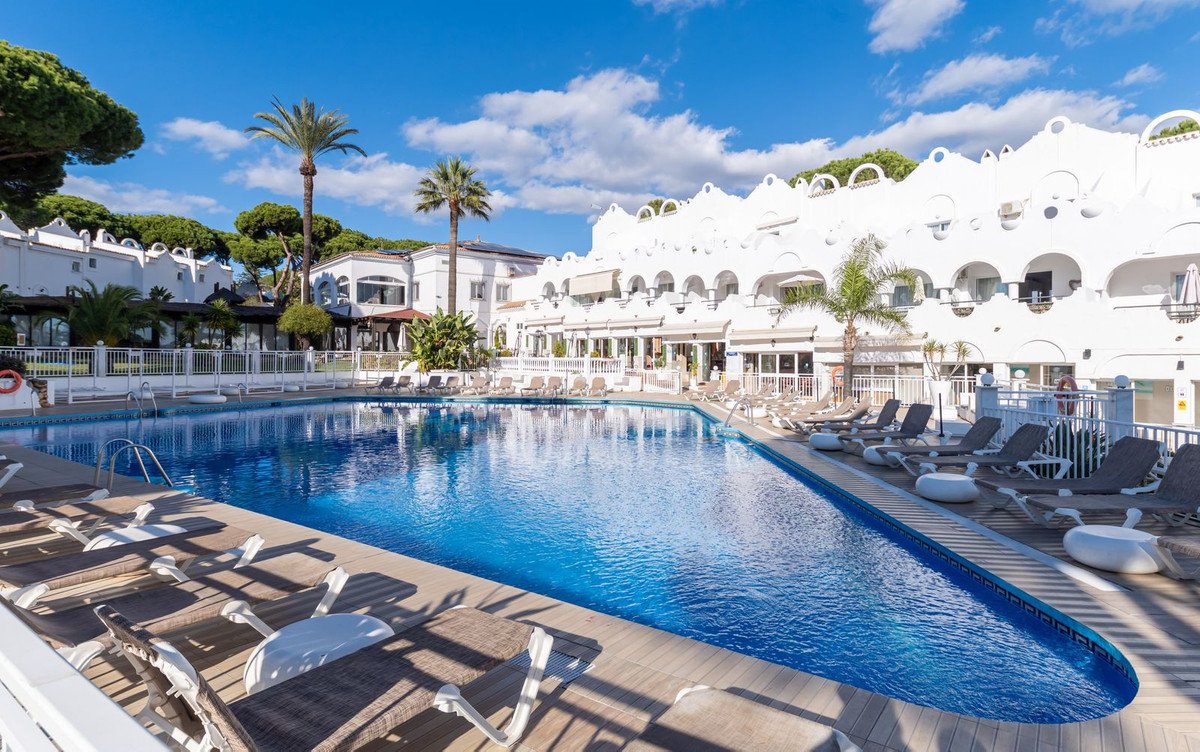 A beautiful outdoor swimming pool area with loungers, palm trees, and white hotel buildings under a blue sky.