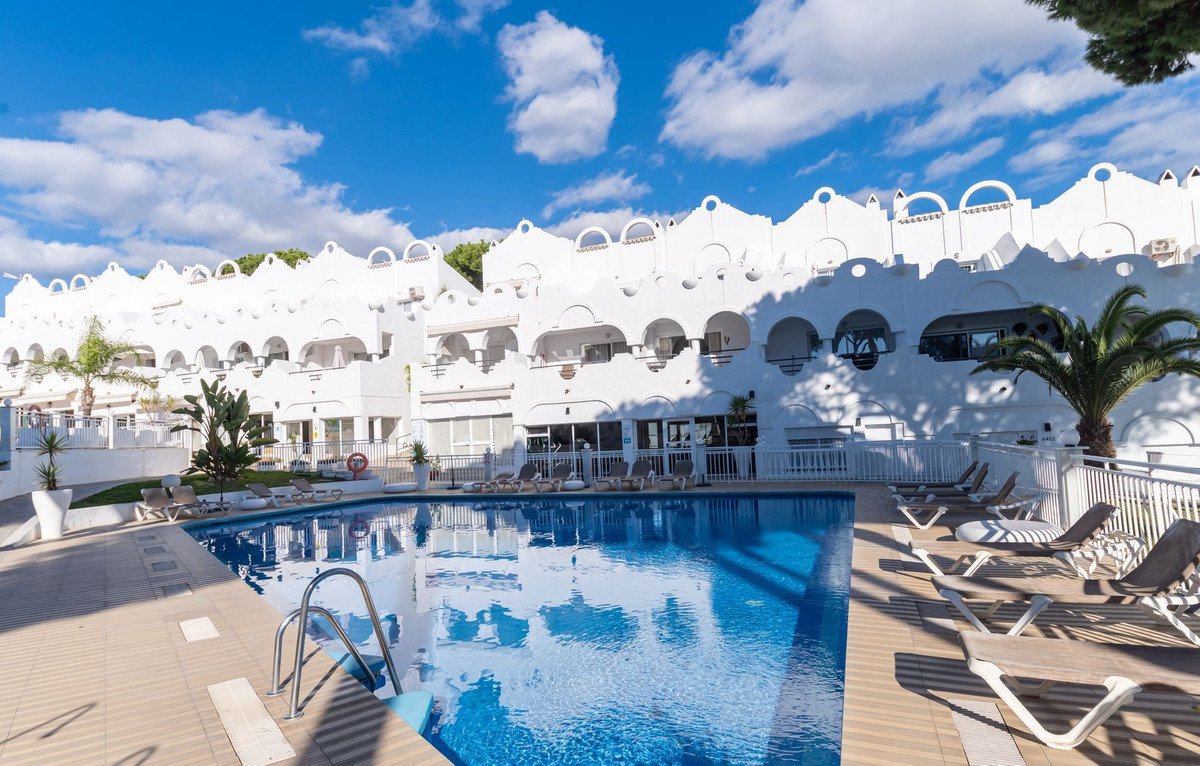 A bright blue swimming pool with lounge chairs and a white resort building under a sunny sky with clouds.