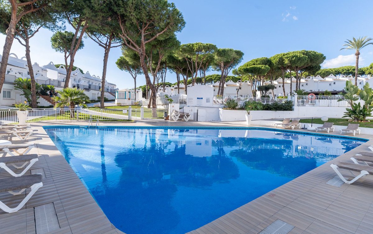 A wide view of a resort swimming pool surrounded by lounge chairs, pine trees, and white apartments under a blue sky.