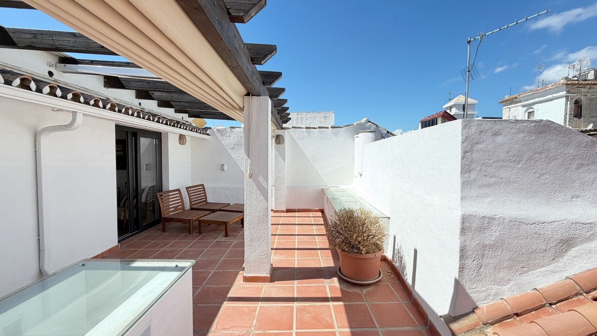 Terrace with wooden chairs, tables, and a pergola, overlooking rooftops under a clear blue sky.