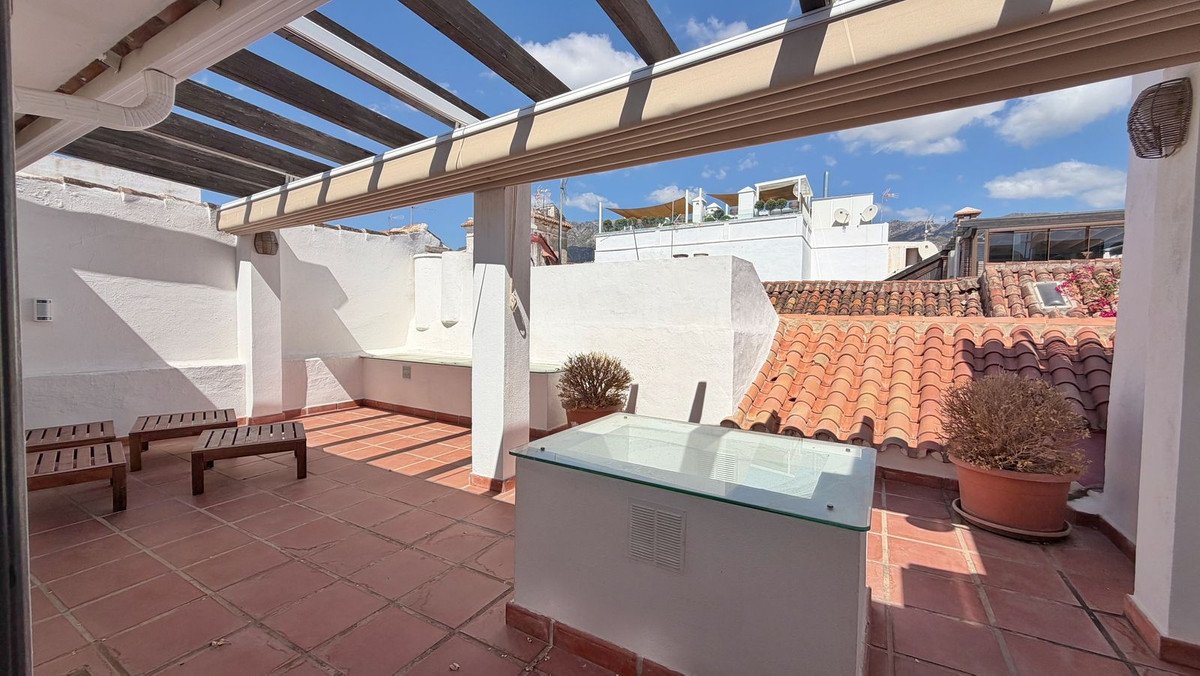 Spacious tiled terrace with wooden benches, pergola, and a glass-covered feature, offering views of rooftops and mountains.