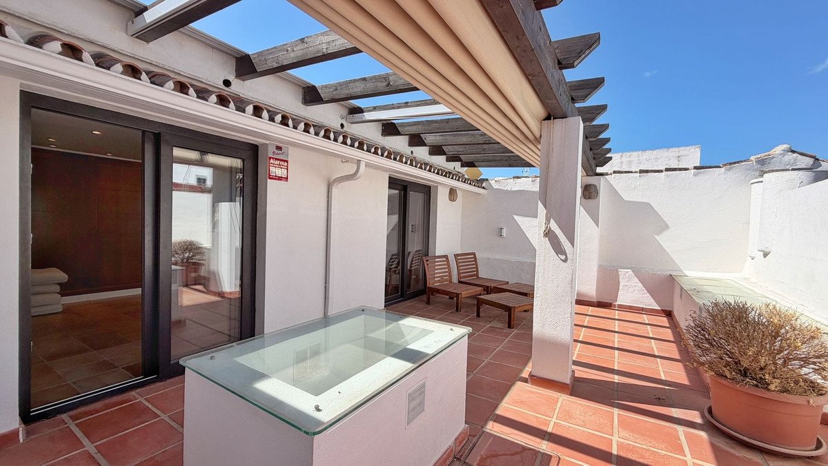 A sunny terrace with terracotta flooring, wooden seating, a glass-top table, and a retractable pergola overhead.