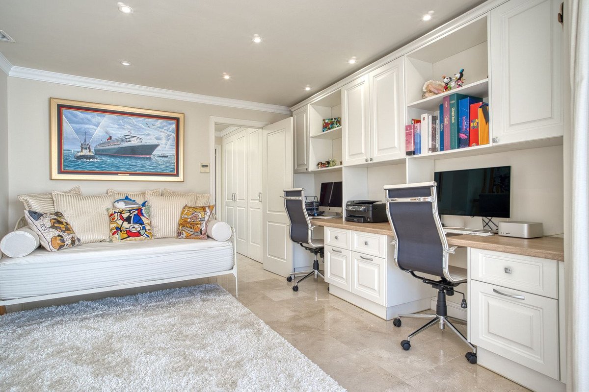 Modern living room featuring a white sofa with colorful pillows, a large ocean painting, and a home office with two desks and computers