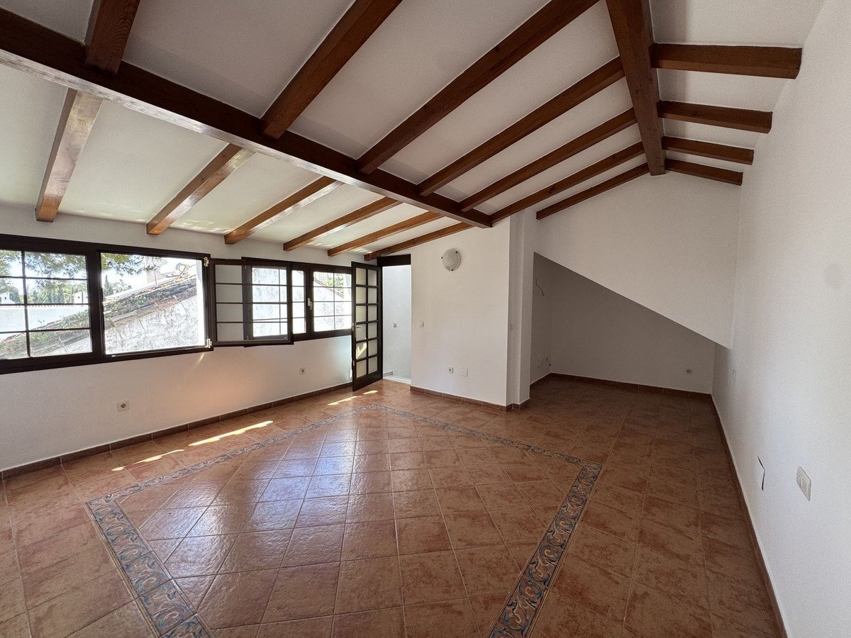 Spacious room with wooden beams, tiled floor, and large windows offering a view of rooftops.