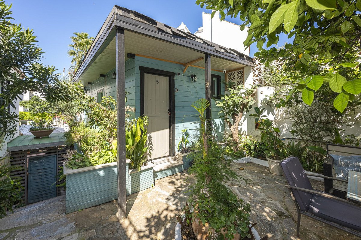A charming blue cottage with a white door, surrounded by lush green plants and trees. Outdoor seating area visible.