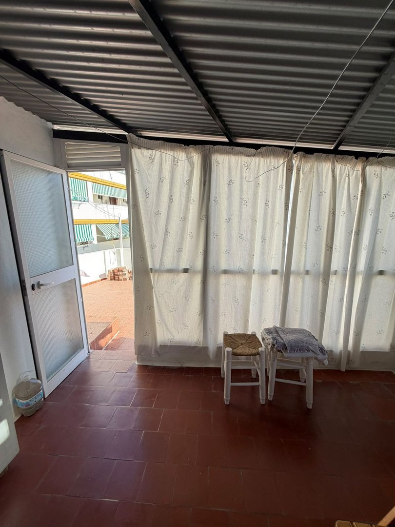 A covered terrace with two small chairs, curtains, and a door leading to an outdoor area with buildings visible in the background.