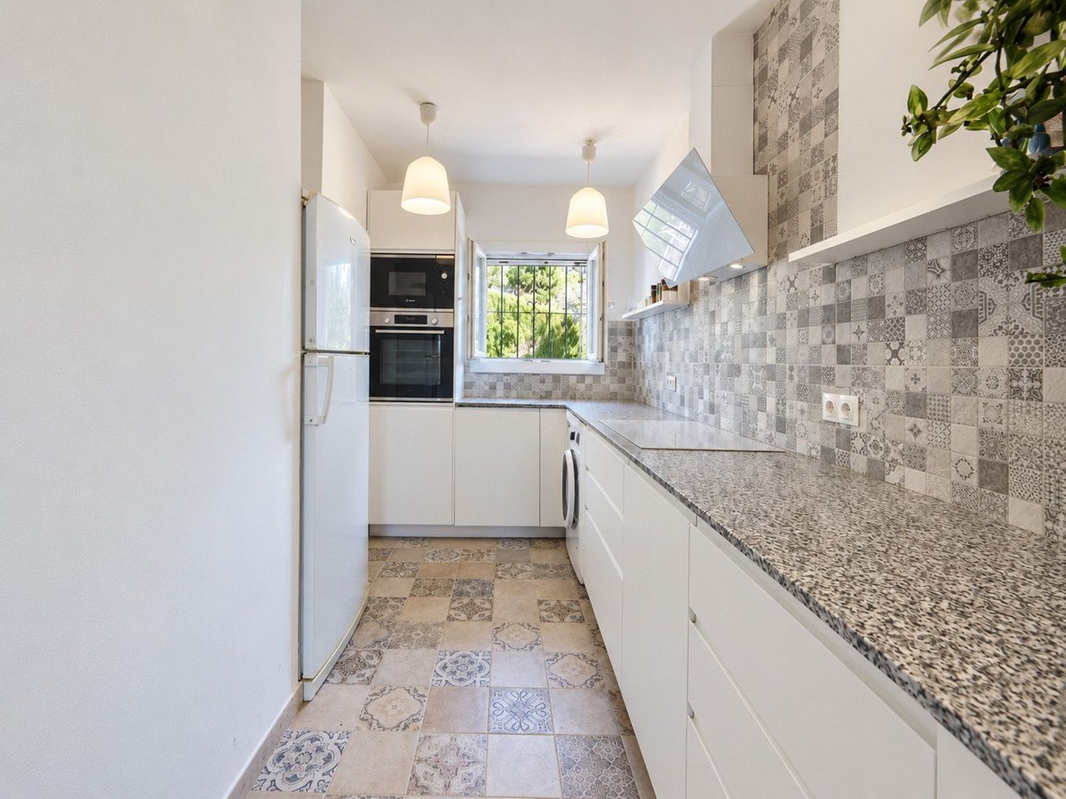 Modern white kitchen with granite countertops, tiled backsplash, oven, microwave, refrigerator, and a window view.