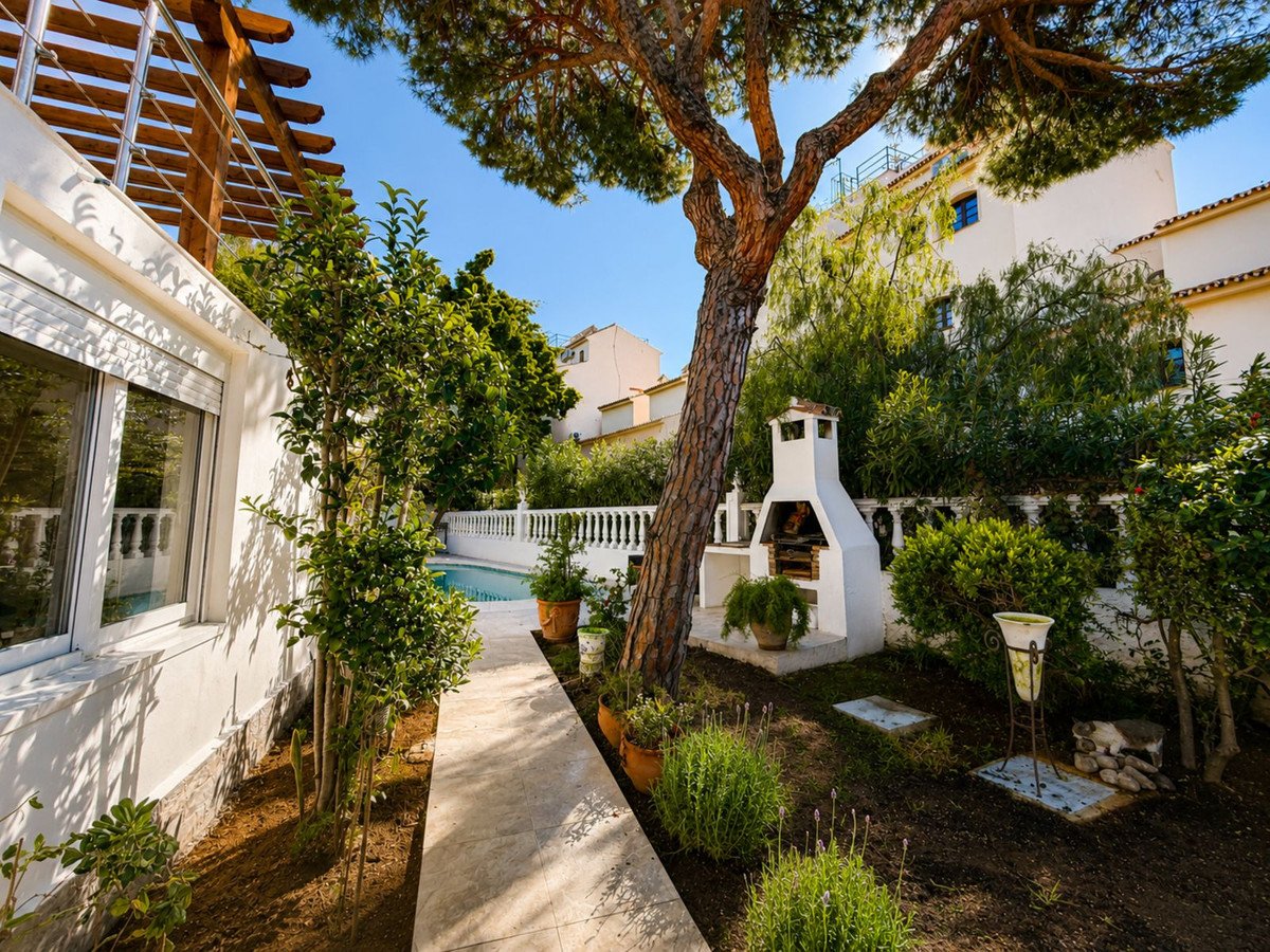 A sunlit backyard with a swimming pool, a built-in barbecue, mature trees, and lush greenery.