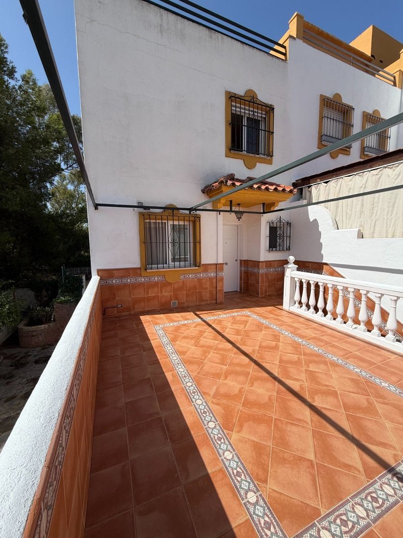 A sunny terrace with terracotta tile flooring and patterned borders, featuring a white balustrade and house facade.