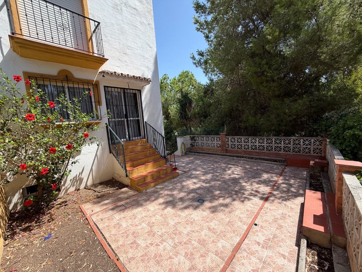 Tiled patio area with stairs leading up to a door and a balcony above, surrounded by lush greenery and trees.