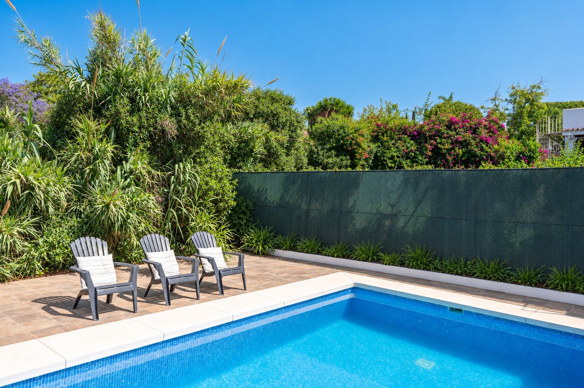 Three chairs with cushions are placed on a tiled patio beside a blue swimming pool with a lush green garden in the background.