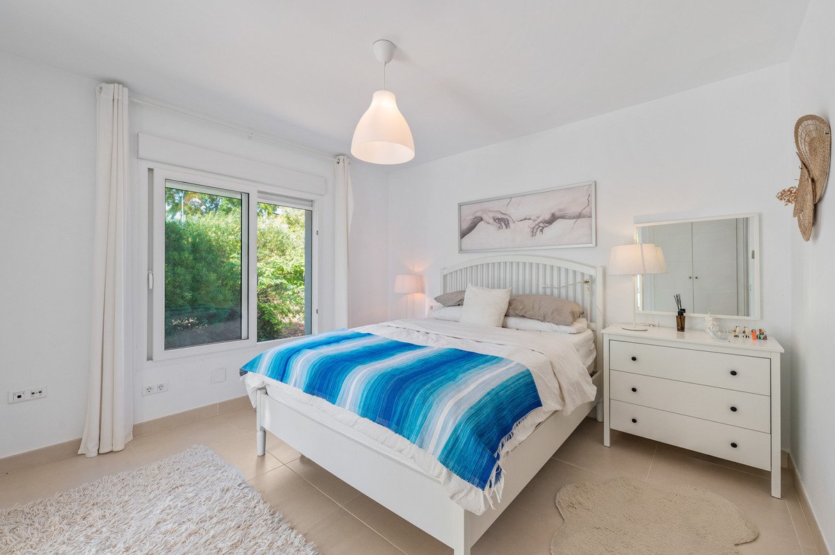 White bedroom with blue striped blanket, large window view of greenery, white furniture, and pendant light.