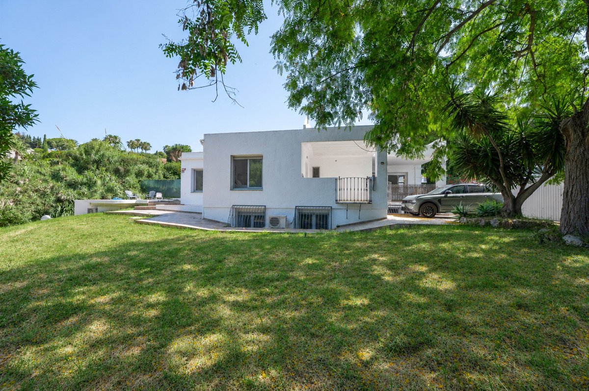 Modern white villa with a green lawn, garden area, and a car parked in the driveway on a sunny day.