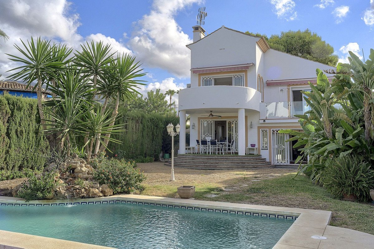 A white villa with a curved balcony, outdoor dining area, and a swimming pool, surrounded by lush greenery and palm trees.