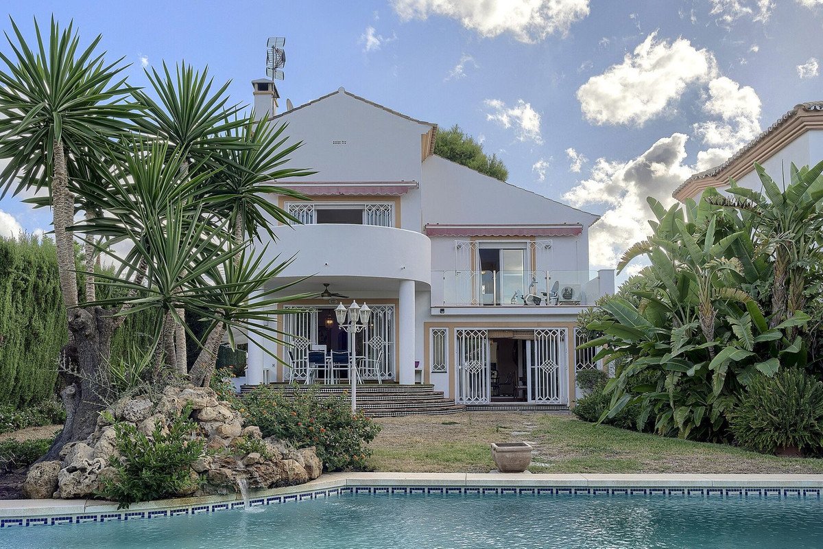 A villa with a swimming pool, palm trees, and lush garden on a sunny day.