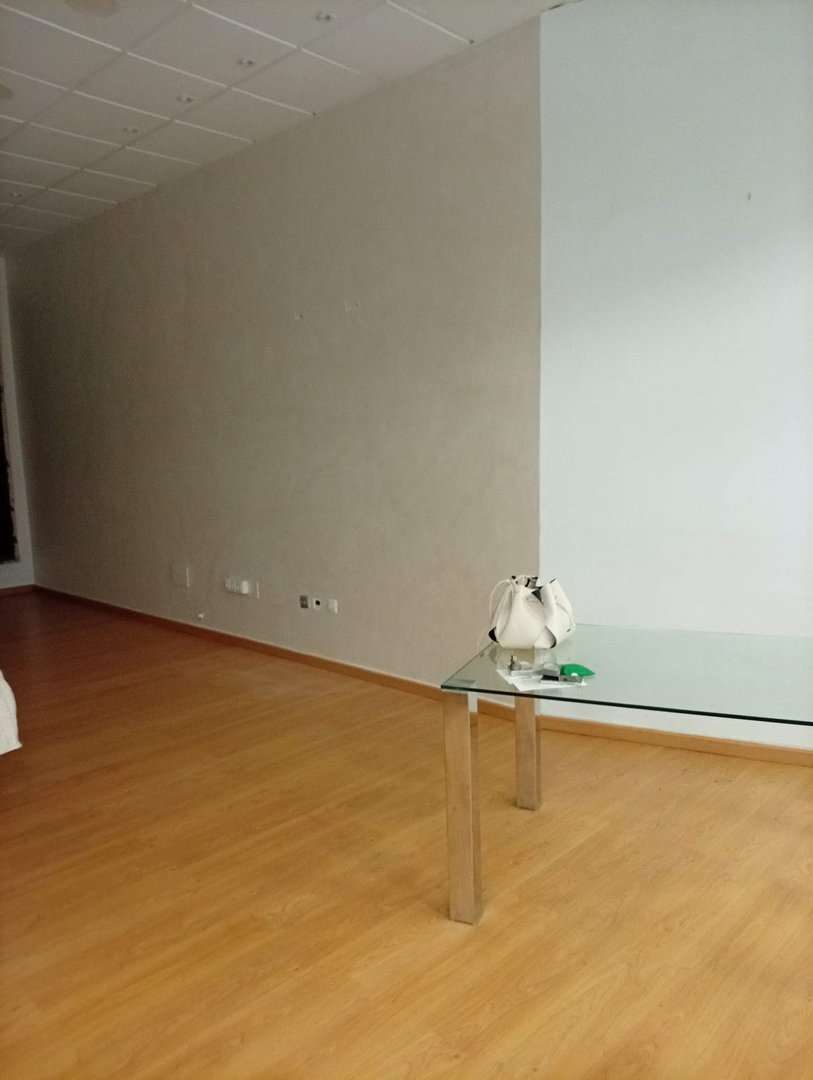 An empty room with light beige walls, wooden floors, and a glass table with a white handbag on it.