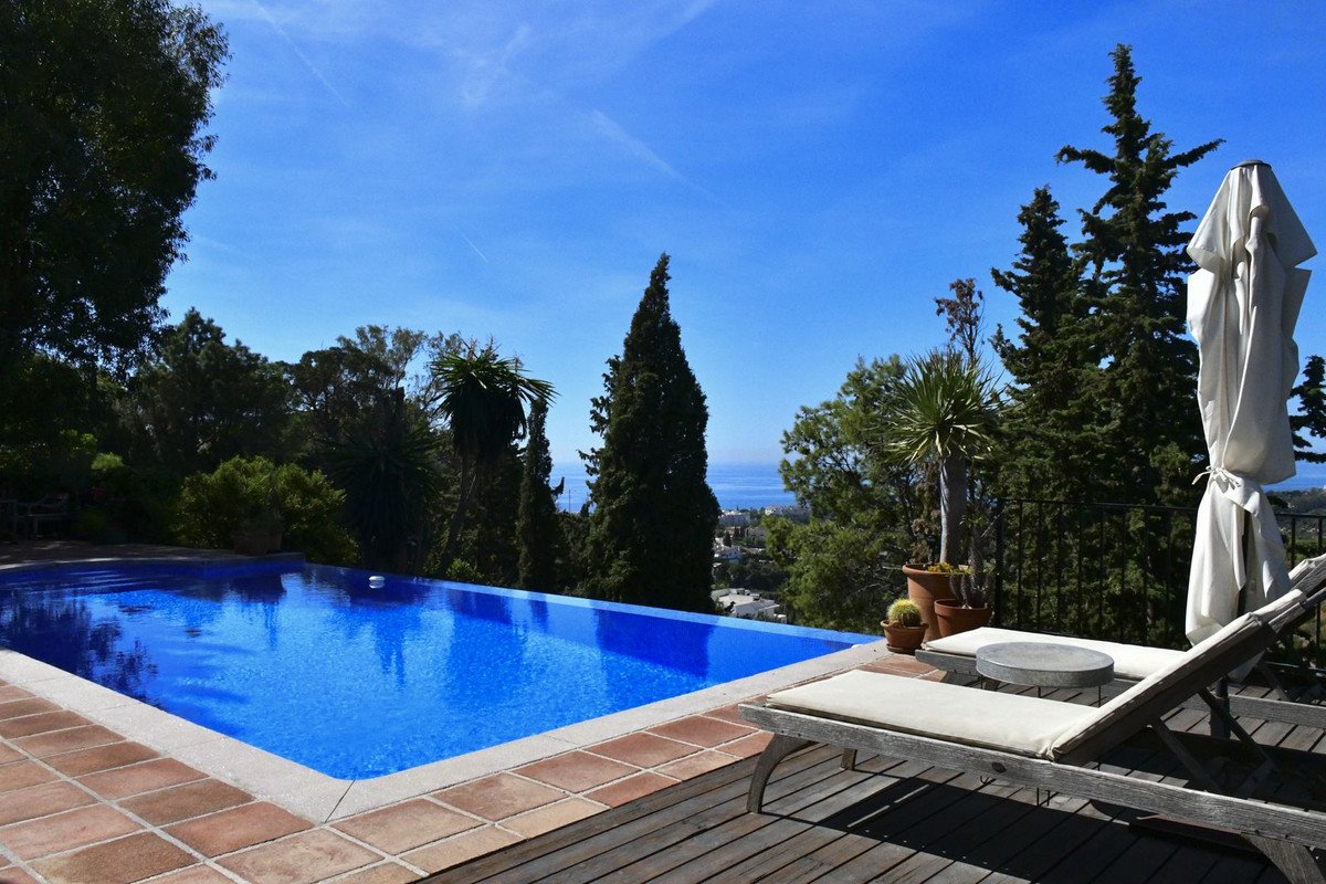 Infinity swimming pool with lounge chairs on a wooden deck, overlooking the sea and surrounded by lush greenery under a clear blue sky.