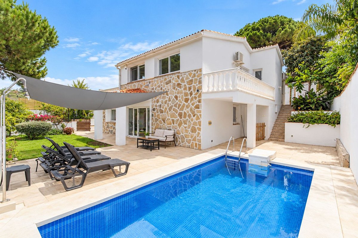 A modern villa with a blue tiled swimming pool, sun loungers, and a shaded patio area on a sunny day.