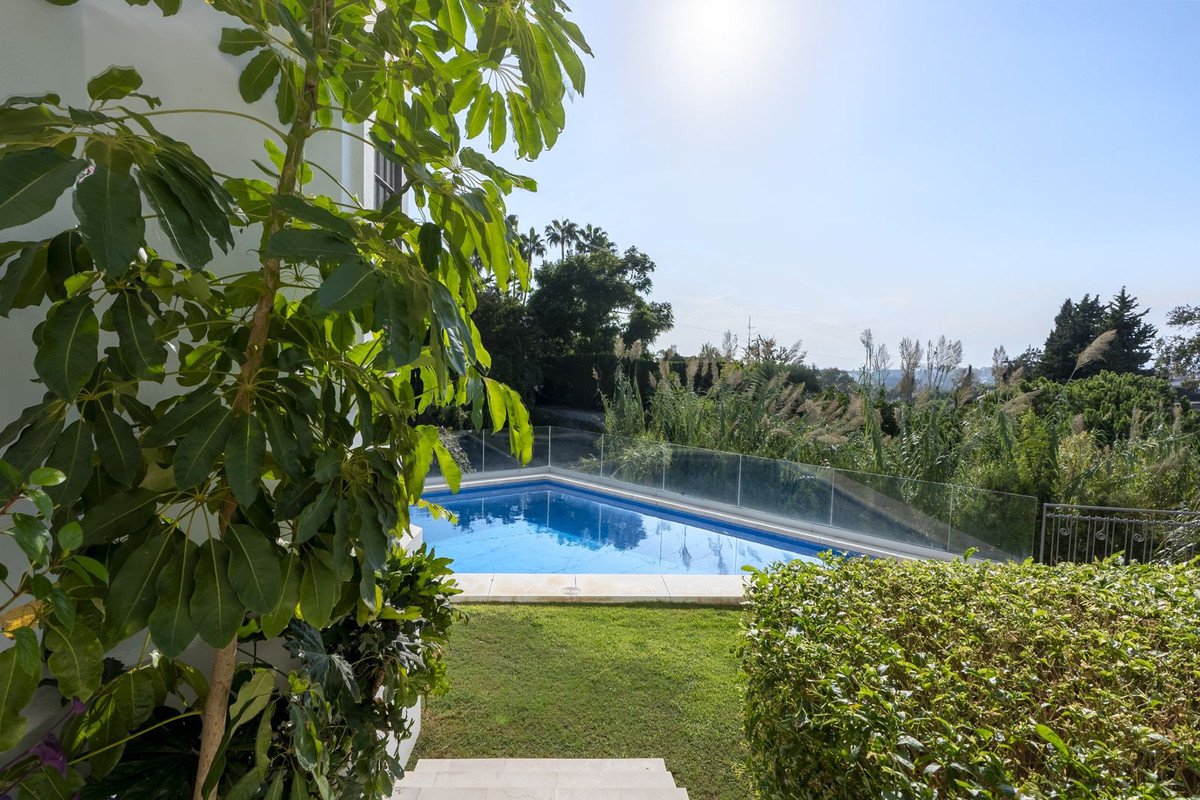 Outdoor swimming pool with a lawn, surrounded by lush greenery and trees, offering a distant view under a clear sky.