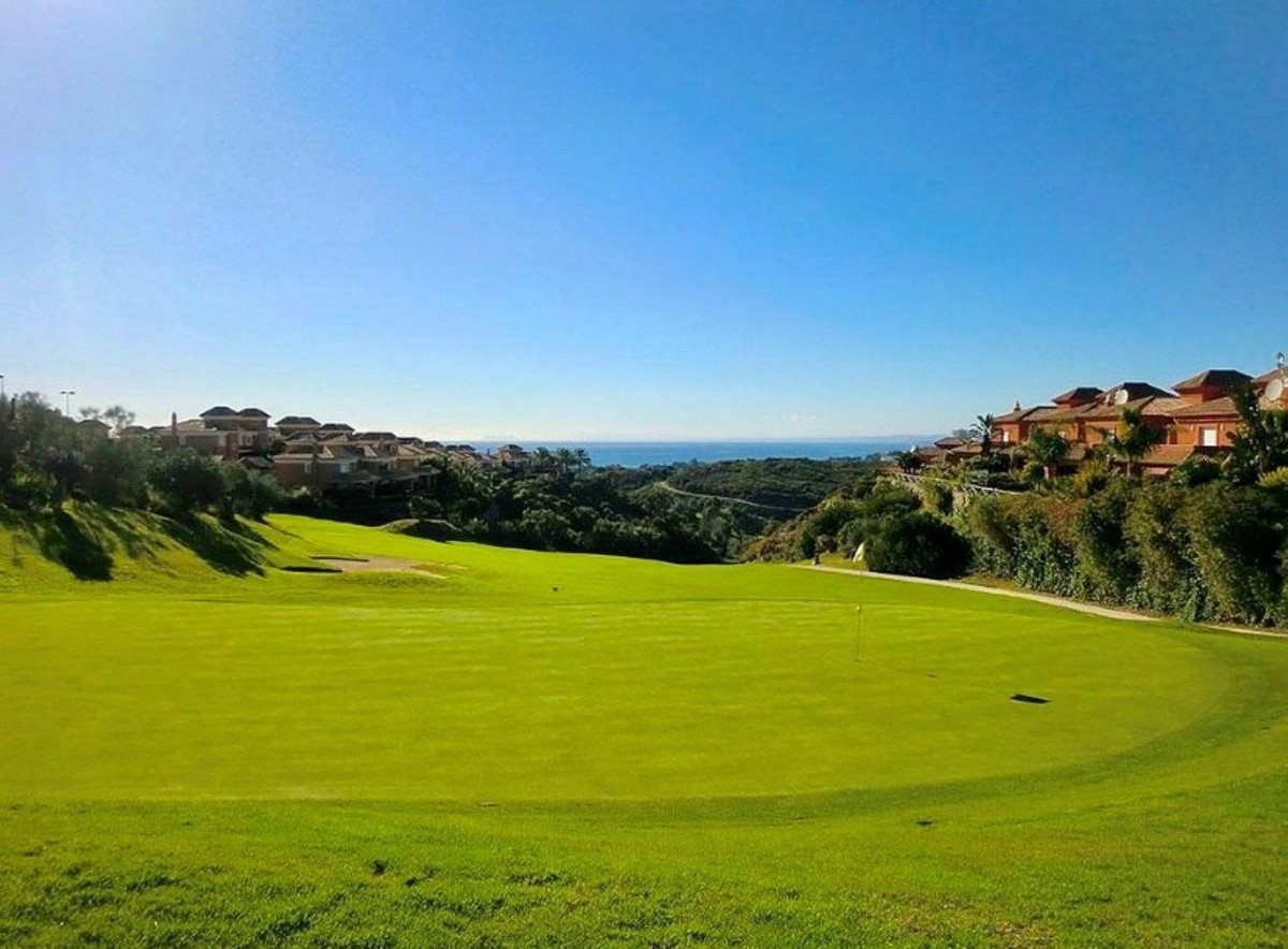 A lush green golf course with houses on a hill and a clear blue sea in the distance under a bright sky.