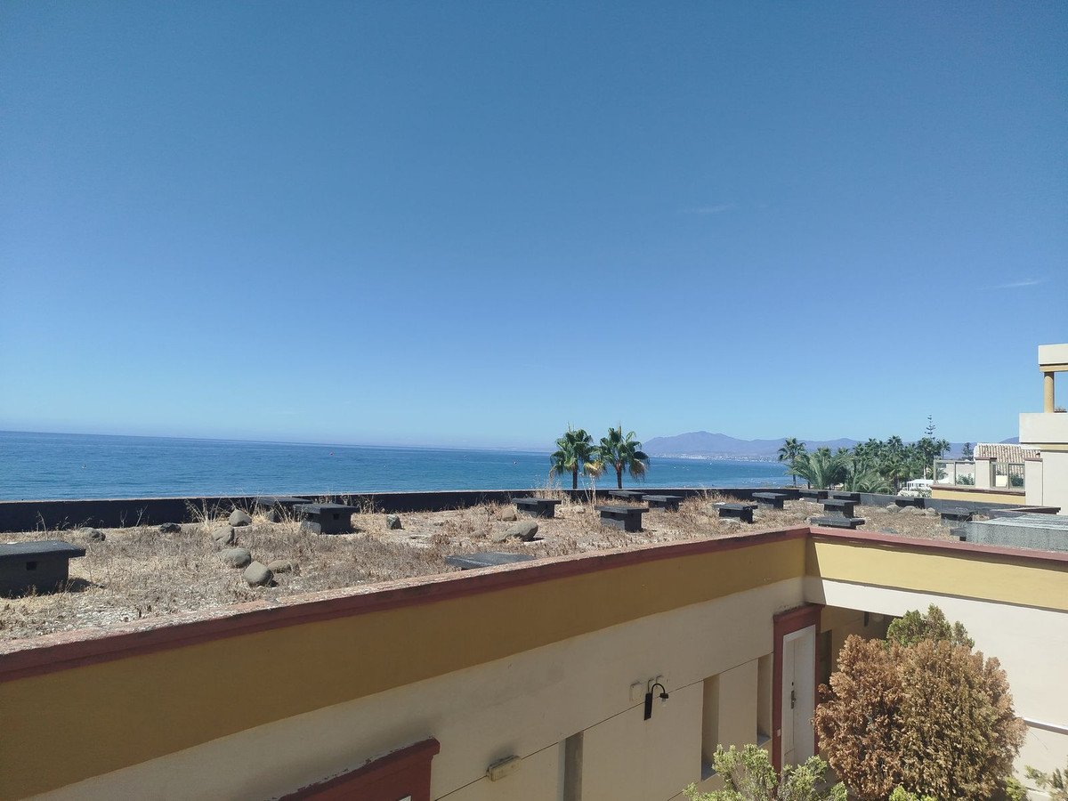 A view from a balcony overlooking the sea with palm trees and distant mountains under a clear blue sky.