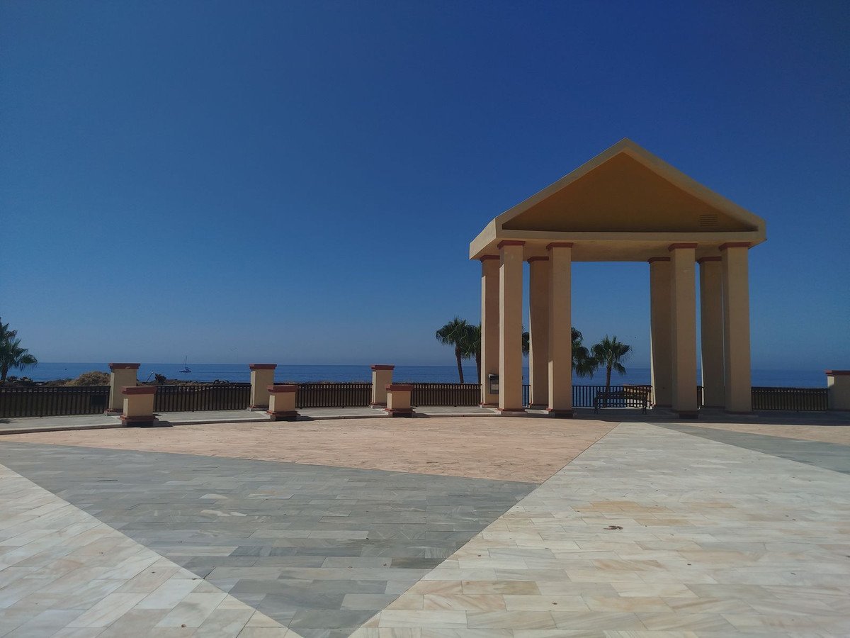 An open-air pavilion with columns overlooks the ocean, with palm trees and a clear blue sky.