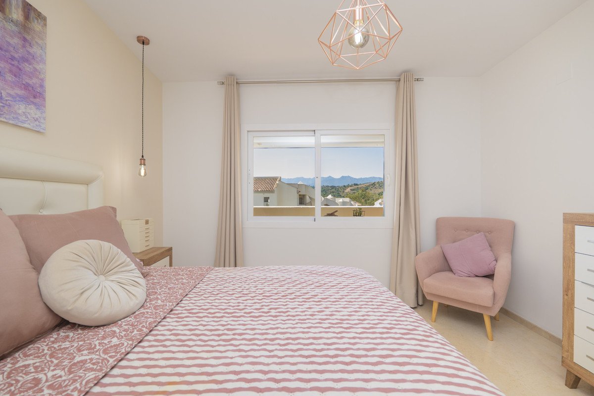 Bedroom with pink accents, a comfortable armchair, and a window offering a beautiful mountain and rooftop view.