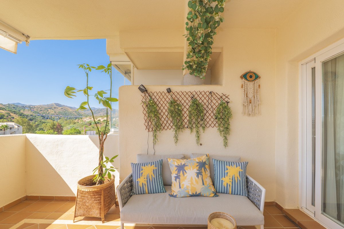 A covered balcony with a sofa, cushions, plants, and a view of green hills under a clear blue sky.