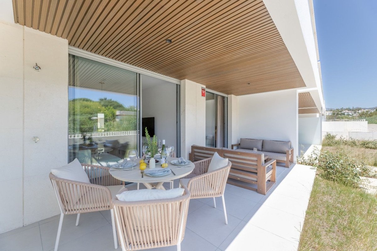 A modern terrace with a dining table, chairs, and a sofa, overlooking lush greenery and a bright blue sky.