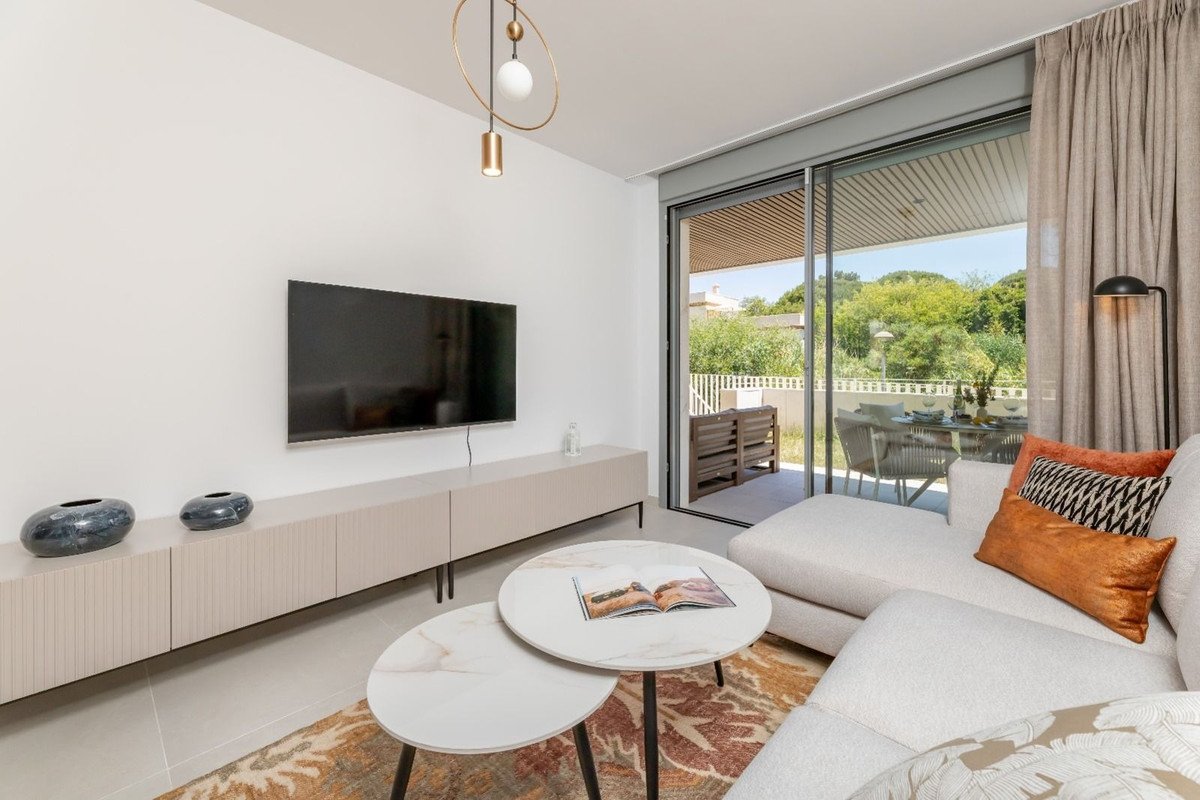 Modern living room with a large TV, sectional sofa, and sliding doors opening to a patio with outdoor dining furniture and greenery.