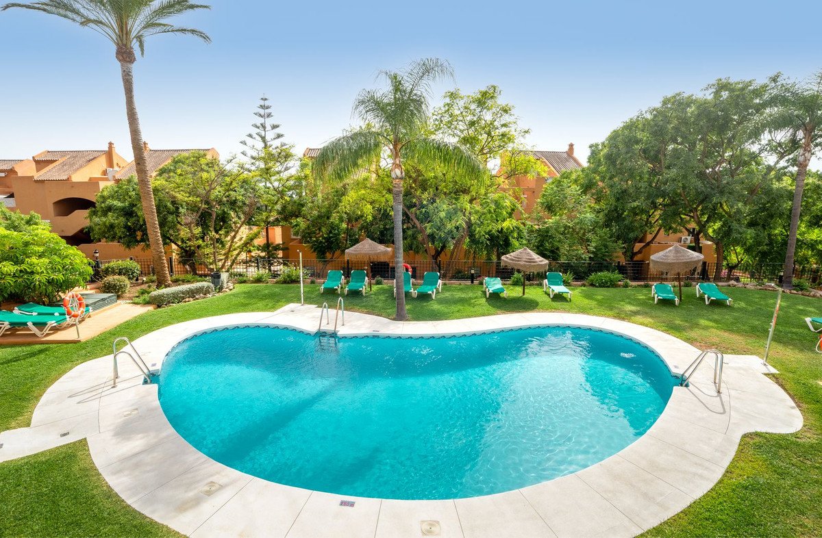 A kidney-shaped swimming pool surrounded by a lush green lawn, sun loungers, palm trees, and residential buildings.
